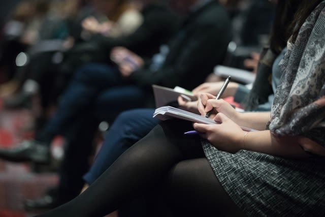Besetzte Stuhlreihen bei einer Konferenz. Zuschauer*innen haben Notizblock auf den Knien und schreiben mit.