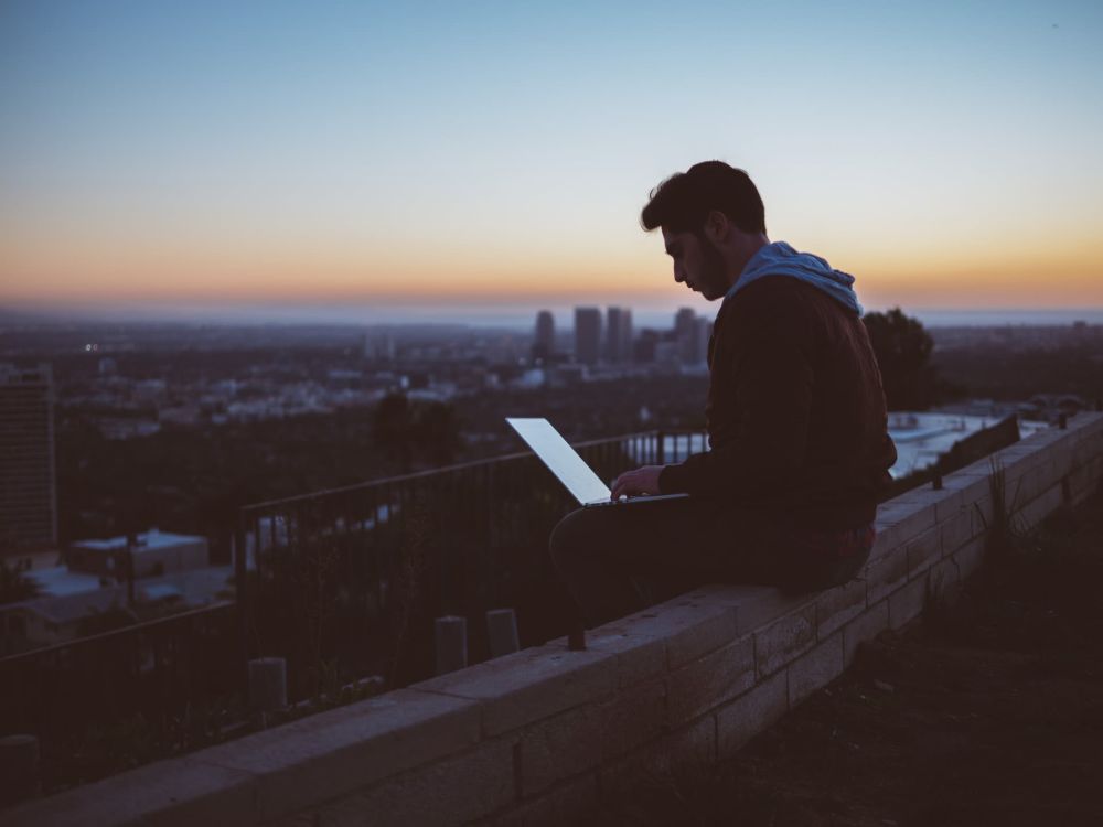 Mann mit Laptop auf dem Schoß sitzt alleine auf einem Dach mit Blick über eine Großstadt