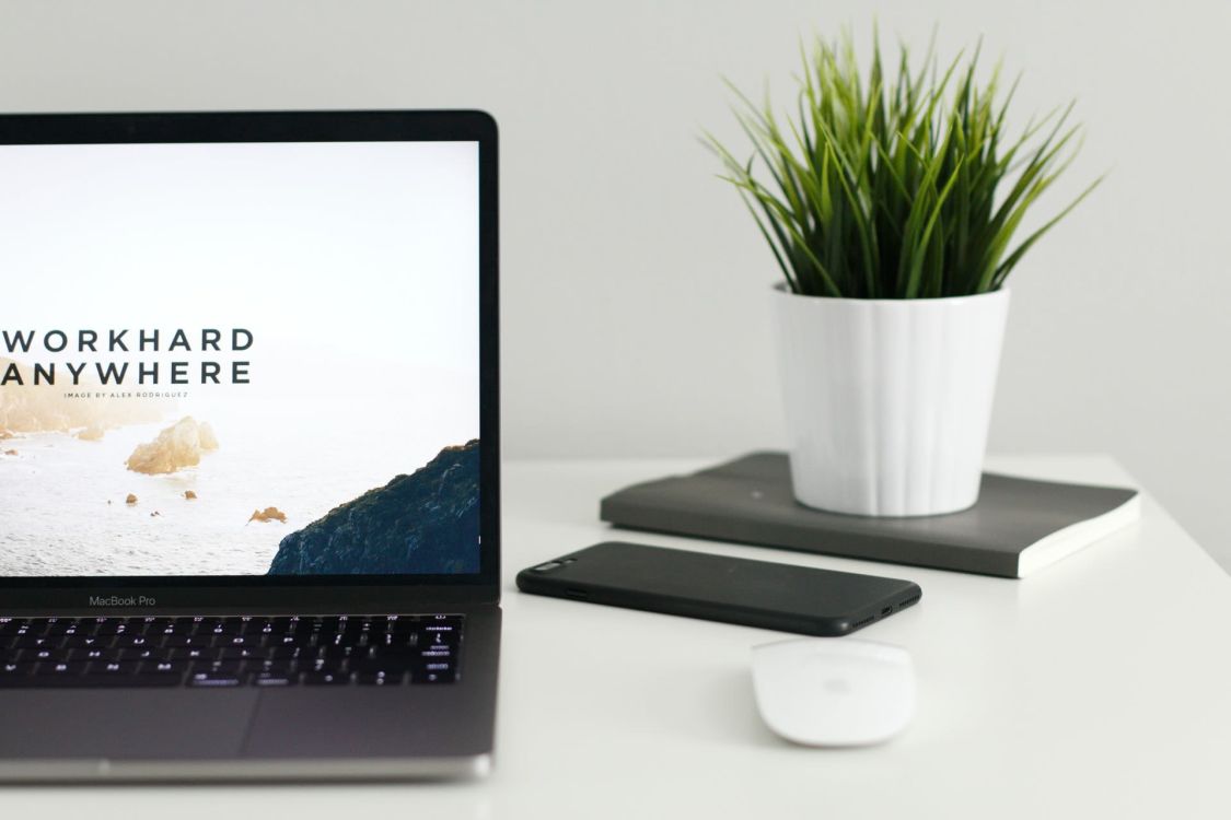 Laptop auf einem Tisch. Eine kabellose Maus, ein Handy und  eine Pflanze daneben. Auf dem Bildschirm steht: "Work hard from anywhere."