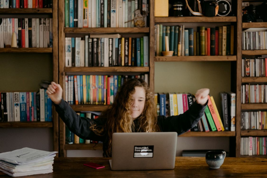M&auml;dchen sitzt in Sieges-Pose vor einem B&uuml;cherregal an einem Schreibtisch vor dem Laptop