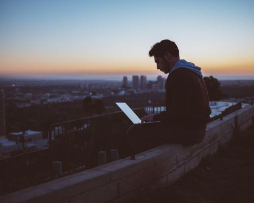 Mann mit Laptop auf dem Schoß sitzt alleine auf einem Dach mit Blick über eine Großstadt
