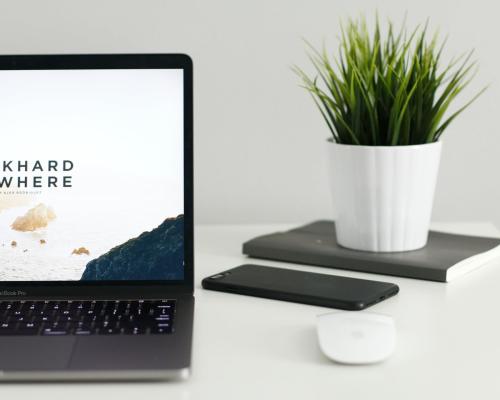 Laptop auf einem Tisch. Eine kabellose Maus, ein Handy und  eine Pflanze daneben. Auf dem Bildschirm steht: "Work hard from anywhere."